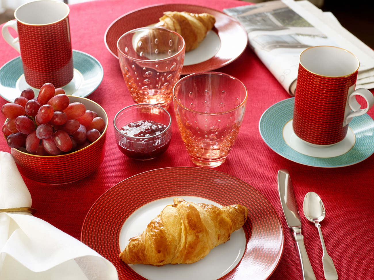 Frühstückstisch mit rotem Geschirr, Croissants, Trauben, Marmelade, Zeitung auf rotem Tischtuch. Fürstenberg Porzellan.