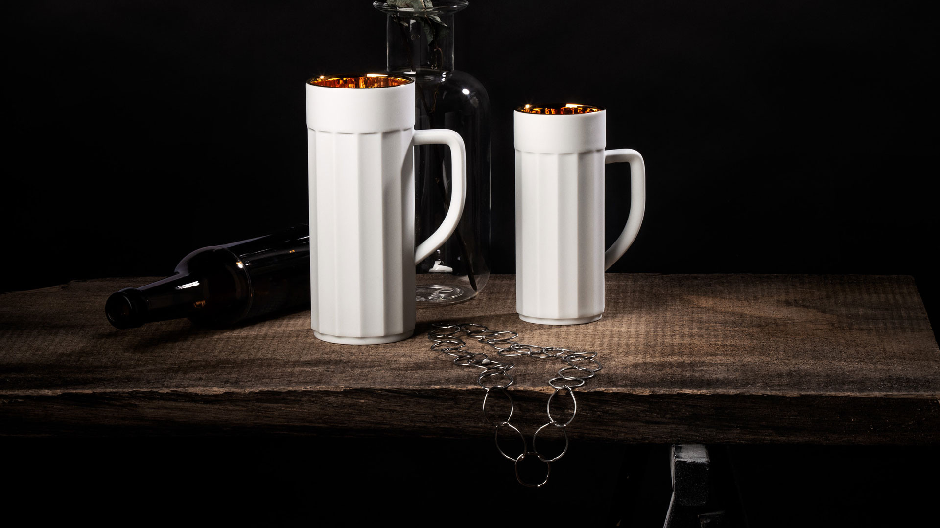 Weiße Porzellanbecher mit goldener Innenfläche stehen auf Holztisch neben Glasflasche. Fürstenberg Porzellan.