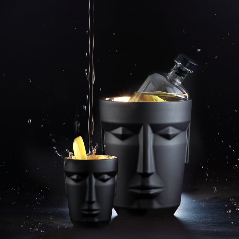 Schwarze Gesichtsvase hält Flasche, daneben Becher mit Zitronenscheibe und Flüssigkeit. Fürstenberg Porzellan.
