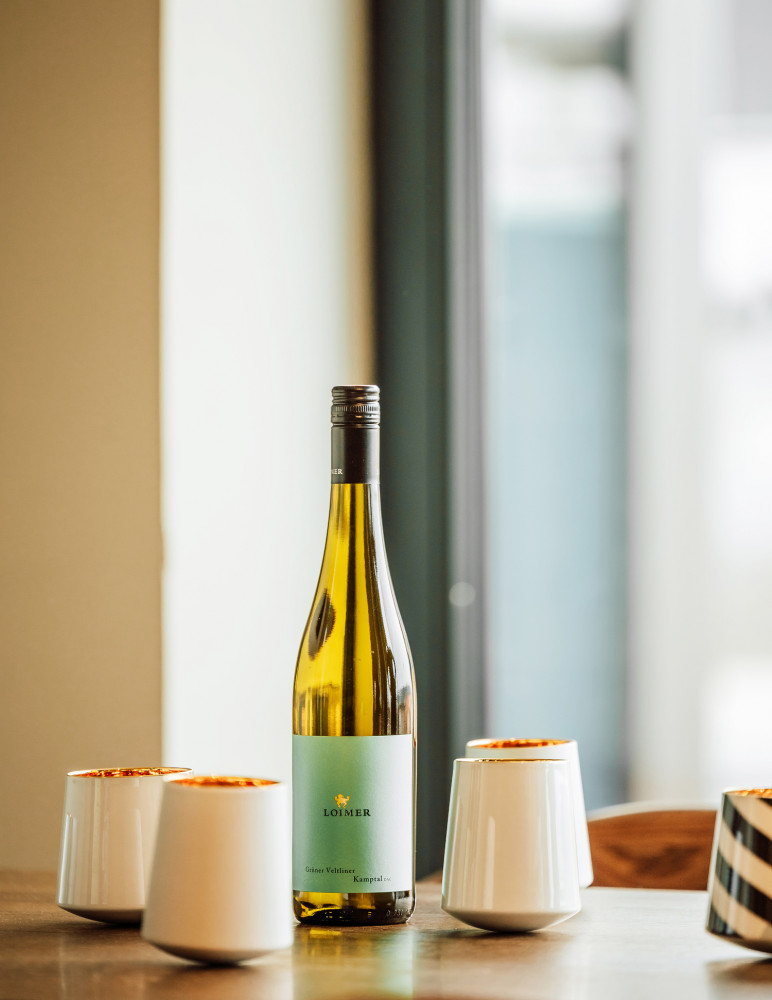 Weinflasche Loimer Grüner Veltliner steht zwischen weißen Bechern auf Holztisch. Fürstenberg Porzellan.