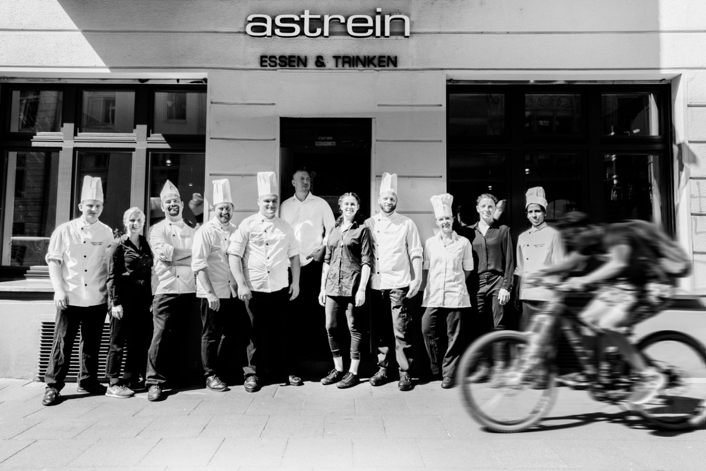 Köche und Kellner posieren vor Restaurant „astrein Essen & Trinken“, Fahrradfahrer vorbeifahrend. Fürstenberg Porzellan.