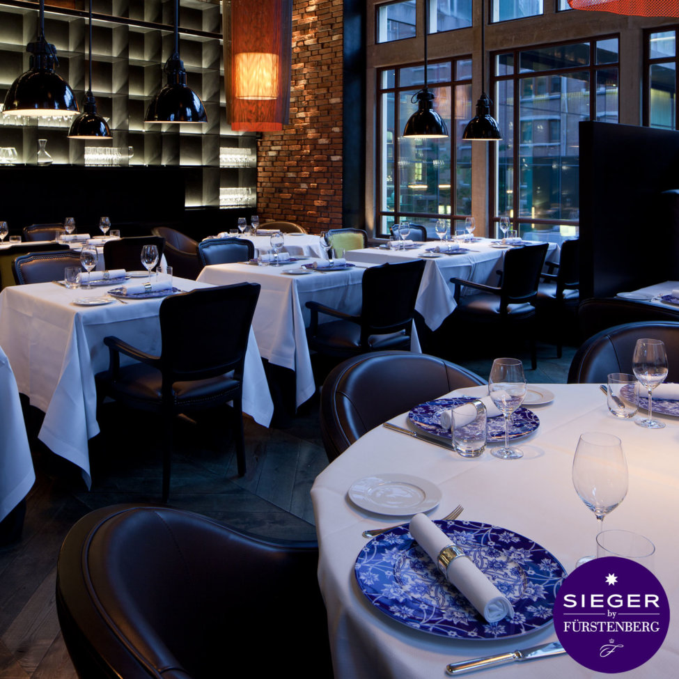 Elegantes Restaurant mit gedeckten Tischen, blauen Tellern und Fenstern; Label: SIEGER Fürstenberg Porzellan.