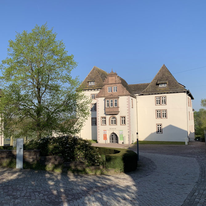 Ein Schlossgebäude mit geteilten Dächern, davor ein großer, grüner Baum. Fürstenberg Porzellan.
