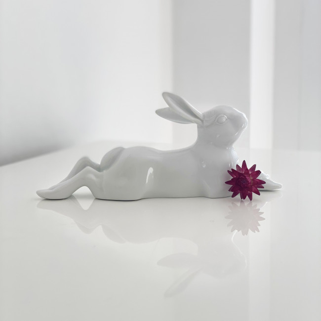 Weiße, liegende Porzellanfigur eines Hasen mit lilafarbener Blume auf weißem Tisch. Fürstenberg Porzellan.