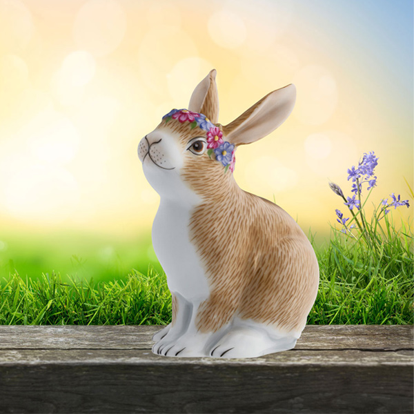 Porzellanfigur Hase, trägt Blumenkranz, sitzt auf Holztisch vor grünem Hintergrund. Fürstenberg Porzellan.