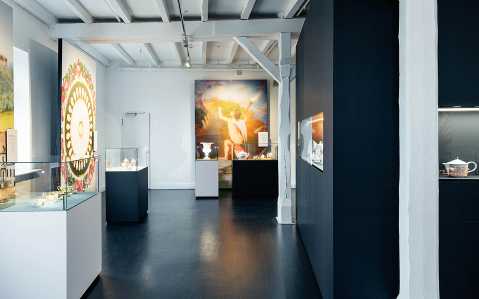 Museumsausstellung mit Vitrinen, beleuchtetem Porzellan, bunten Wandbildern; helle, moderne Umgebung. Fürstenberg Porzellan.