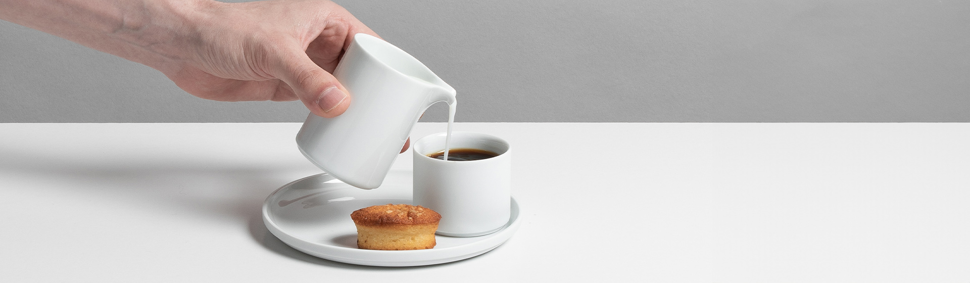 Hand gießt Milch in weißen Kaffee neben Muffin auf weißem Teller. Fürstenberg Porzellan.