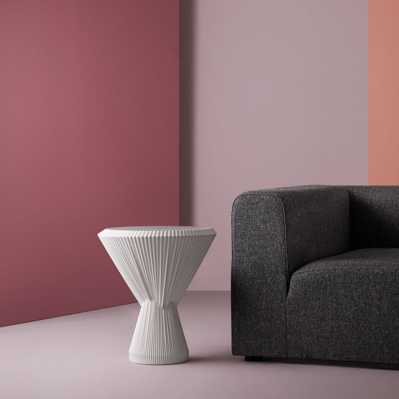 Weißer, gefalteter Beistelltisch vor grauem Sofa auf rosa Hintergrund. Fürstenberg Porzellan.