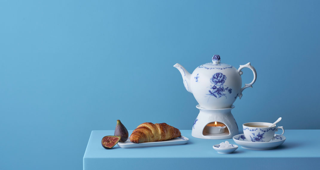 Teekanne erhitzt sich über Kerze neben Croissant und Feige auf Tisch. Fürstenberg Porzellan.