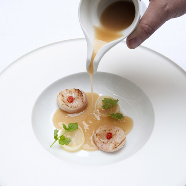 Jakobsmuscheln mit Sauce in weißem Teller serviert, Hand gießt Sauce. Fürstenberg Porzellan.
