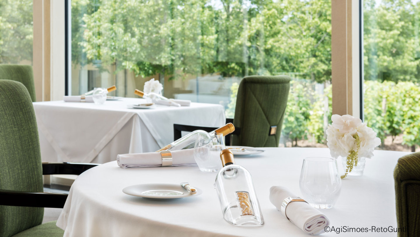 Gedeckter Tisch mit weißen Tischdecken, grünen Stühlen und Blumenarrangement vor Fenster. Fürstenberg Porzellan.