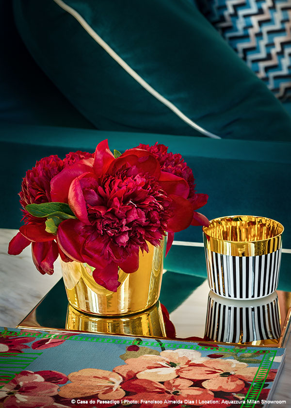 Goldene Vase mit roten Pfingstrosen, auf verspiegeltem Tisch vor blauem Sofa. Fürstenberg Porzellan.