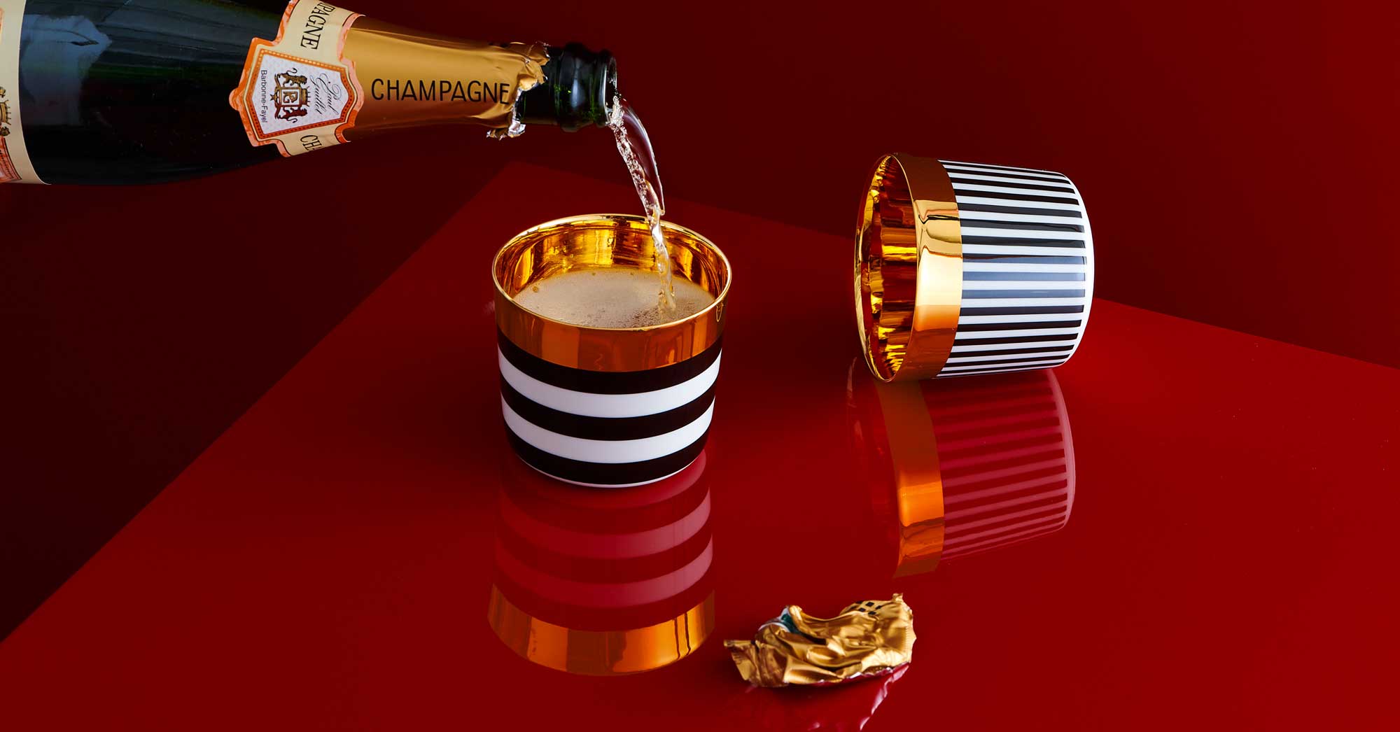Champagnerflasche schenkt in gestreiftes, goldverziertes Glas auf rotem Tisch ein. Fürstenberg Porzellan.