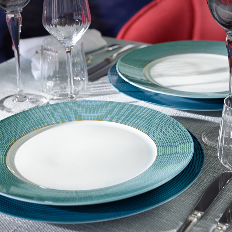 Teller mit blau-grünem Rand auf gedecktem Tisch, neben Weingläsern und Besteck. Fürstenberg Porzellan.