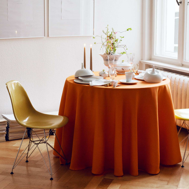 Runder Tisch mit orangefarbener Tischdecke, gedeckt mit Porzellan, Kerzen und Vase. Fürstenberg Porzellan.