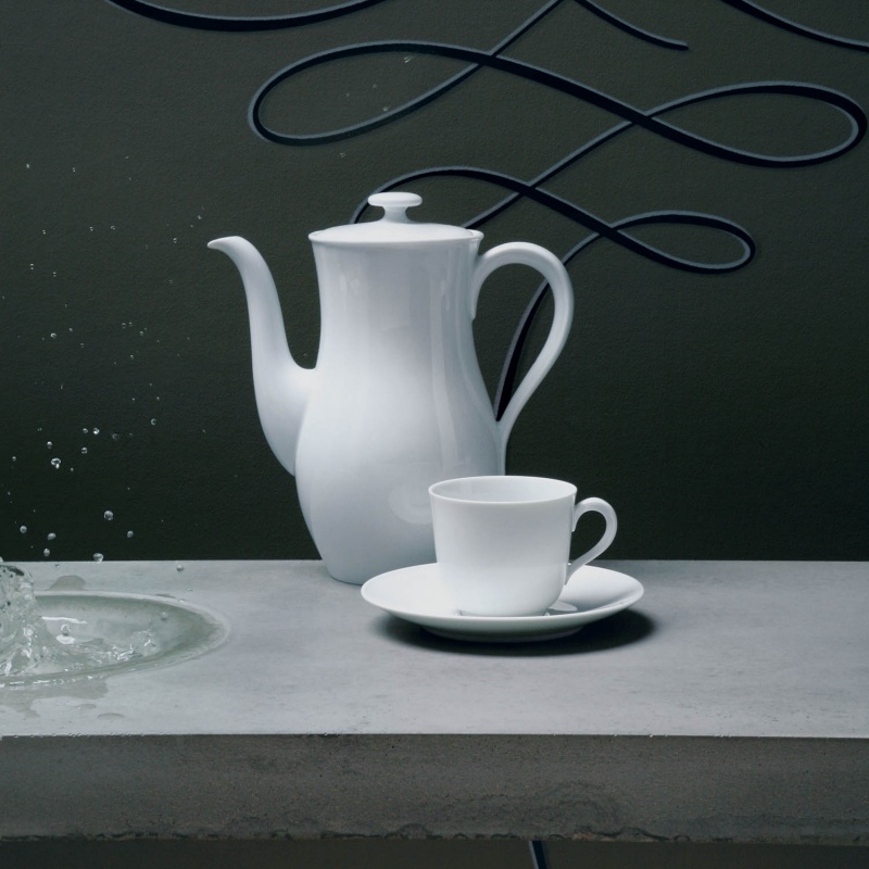 Weiße Kaffeekanne und Tasse auf grauem Tisch, Wasser spritzt daneben. Fürstenberg Porzellan.